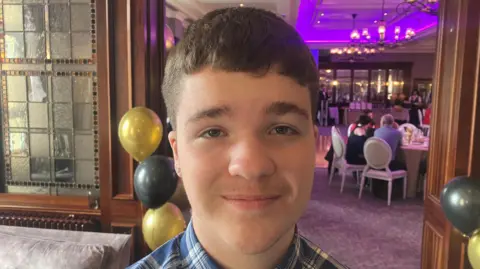 Jamie Harte is standing in an events space with different tables set out and a blue projector screen. A sofa and chairs are visible to his left. He has short brown hair and is wearing a blue chequered shirt.