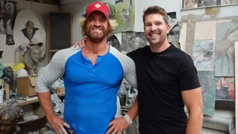Nick Elphick wears a blue and grey long-sleeved top and red baseball hat. He is standing next to Darryn Frost, who has his arm around him. He is wearing a black T-shirt. They are both smiling at the camera and standing in an art studio, with paintings and small sculptures in the background.