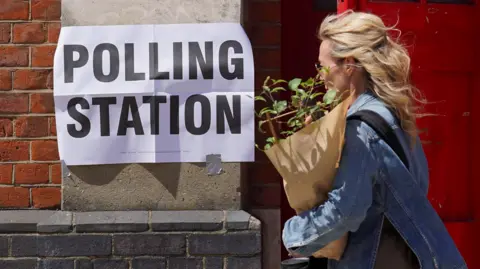 A woman carrying a plant wrapped in brown paper walks past a building with a polling station sign taped to it