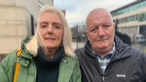 Brother and sister Maggie and John Crawford. Maggie has blonde hair, wearing a green coat, navy jumper and carrying a yellow bag. John is bald and wearing a black jacket and blue and white shirt.