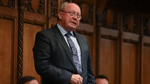 PA Media Jim Allister is speaking in the House of Commons. He is standing up with a brown wood panelled wall behind him. He is wearing glasses, a grey suit, white shirt and floral tie. 