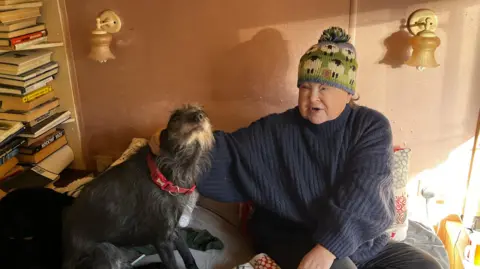 A woman in a woolly jumper and knitted hat sits on the bed of a mobile home with a grey dog, in the background are piles of books and oterh belongings. 