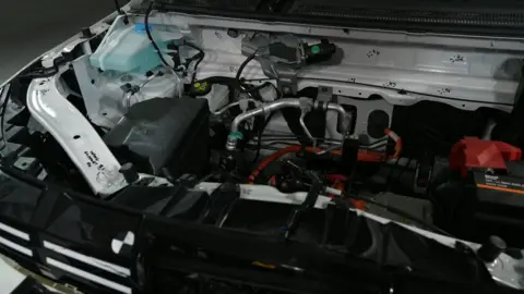 The inside of an EV under the bonnet. The components and the damage to them are not immediately obvious to a layperson. There are metal pipes, rods, wires, and plastic pieces of kit.