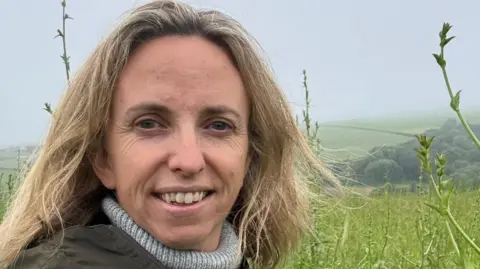Cathy Case A picture of Cathy smiling at the camera. She has blonde hair and is wearing a grey jumper and green coat. She is stood in the middle of a field looking over a number of trees and fields.