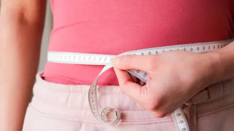 BBC A woman in a pink top with a tape measure around her waist