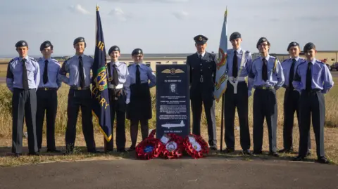A group of 10 military cadets in uniform stand at either side of a black gravestone-type memorial for Flight Captain Susan Shaw on a tarmacked spot on an airfield. Several poppy wreaths lay at the foot of the memorial. Sun-baked yellow grass can be seen behind them. 