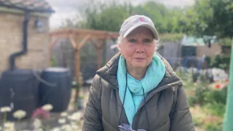Mousumi Bakshi/BBC Eleanor Seers in her garden in a green coat, green scarf and and grey cap.