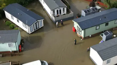 PA Media Another drone image of flooding in Cogenhoe, this time showing five static caravans which are either white or light green in colour. A few people are standing in brown water that, in terms of depth, is just under their knees. The water is totally surrounding each caravan.