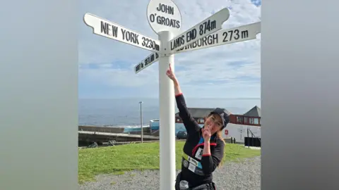 Avril Langard-Tang stands next to a sign on the coast and points to the top which reads "John O'Groats". The sign also directs towards "New York 3,230m", "Edinburgh 273m" and "Lands End 874m". She has her finger on her lips in reference to her challenge, which is called "SilentSteps". You can see the sea behind her.