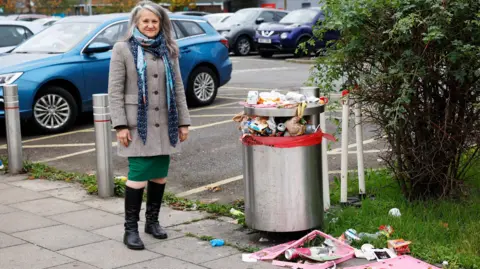 Olivia is stood facing the camera, next to an overflowing street bin and litter strewn all over the payment. 