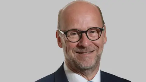 Fareham Borough Council Simon Martin has a bald head, grey stubble and black circular-framed glasses. He is stood in front of a grey wall.