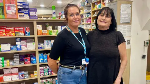 Two women stand in a pharmacy, one with a blue top and jeans, the other with a black dress