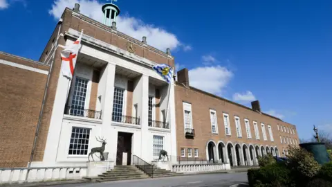 The outside of Hertfordshire County Council's headquarters in Hertford, showing a grand Georgian-style brick building with flags blowing in the wind. Two deer statues are positioned either side of the staircase to the building's entrance. 