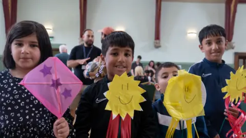 Rochdale Council handout Four young children - one girl (left) and three boys - hold up artwork they have made at a workshop inside Rochdale Town Hall. The girl has a pink paper diamond with three blue stars on it while the boys all have yellow smiley faces. The children are all smiling. 