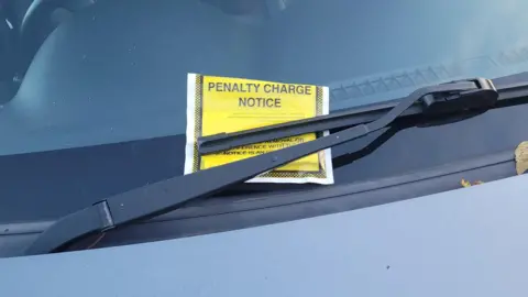 A close-up of a yellow penalty charge notice placed behind the windscreen wiper of a car.