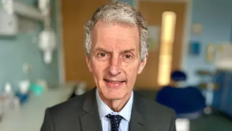 Martin Giles/BBC UEA vice‑chancellor Prof David Maguire wearing a grey suit blue check shirt and tie looking at the camera.