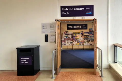 PA Entrance to the Poole Hub and Library, with open wooden double doors leading into a library lobby. A large purple sign above the doorway reads “Hub and Library Poole” with the BCP Council logo. Inside, a “Welcome” sign hangs above displays of brochures and leaflets. To the left of the entrance is a black return box labeled “Returned items and post.” The space has blue carpeting inside the library, light-colored walls, and a tiled floor in the foreground.