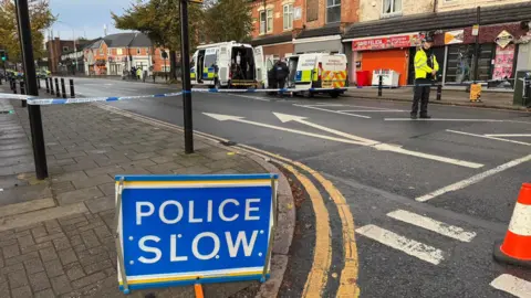 Leicester Media Online A police slow sign in front of a taped off road with a police officer standing at the cordon and two police vans in the background