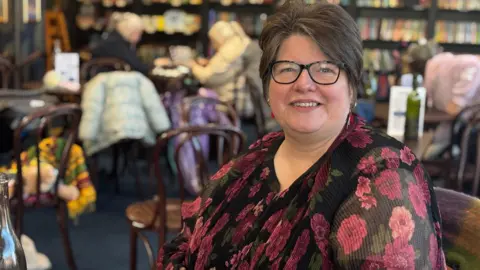 Claire Hamilton/BBC Claire Morgans is wearing a black dress with purples flowers on it. She has brown hair and black spectacles.