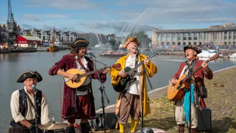 Bristol Harbour Festival Piratitude, a male musical group dressed up as pirates. They are all playing instruments and singing by the waterfront in Bristol.