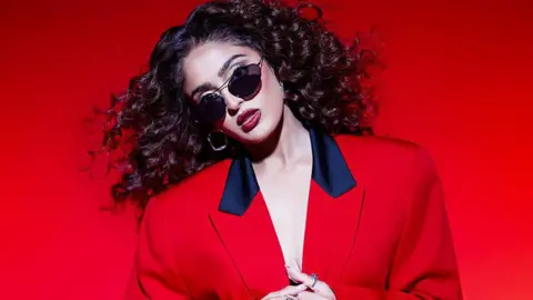 Sequoia Emmanuelle Mandeep Dhillon, a woman wearing sunglasses and a red blazer, in front of a red background.