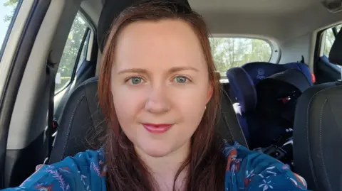 Rachel has taken a selfie photo in her car. She has shoulder length brunette hair and is wearing a blue blouse top. 