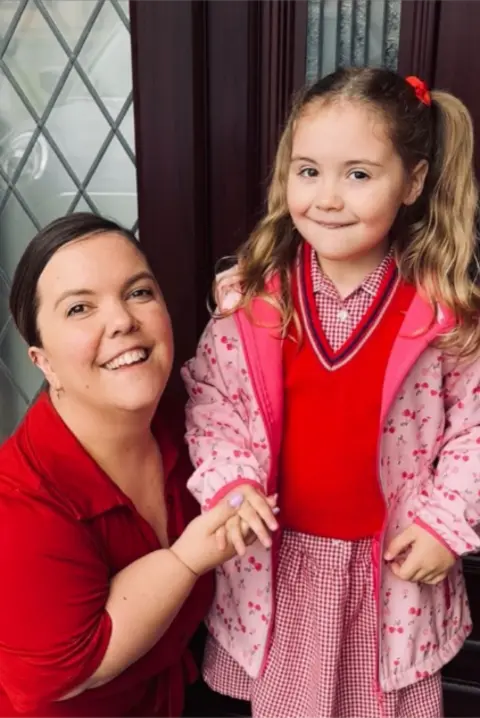 Family Fund Hanna is kneeling next to Darcy a the inside of a front door with a decorative lead frame window. Darcy has long blonde pig tail hair and is smiling for the cameral with a pink winter jacket and red jumper. Mum has long tied back brown hair and is smiling