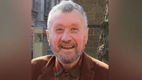 A man with grey hair and a grey beard smiling at the camera. He is wearing a brown jacket and a red and black checked shirt.