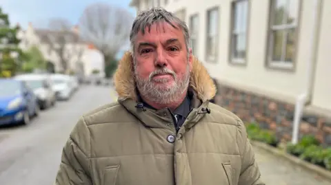 MP Peter Roffey stands before the royal court of Guernsey. It has a gray beard and average gray hair. He wears a green jacket swollen with frills on the hood.