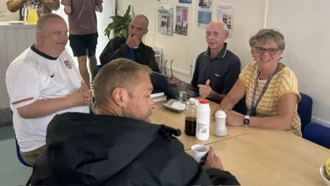 BBC/Melanie James Five people seated around a table in a community café or meeting space. Drinks, cups, and condiment containers are on the table. The room has white walls with posters, noticeboards, and leaflet displays, while additional people are visible near a serving counter in the background.