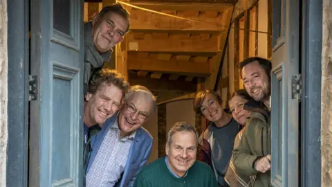 A group of seven middle-aged and older people, five men and two women, are peering around blue double doors as if they have just answered the doorbell, and are smiling at the camera.