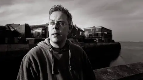 Nick Quantrill. A moody black and white photograph taken outside with a boat in the background.  He wears a hoodie and glasses.