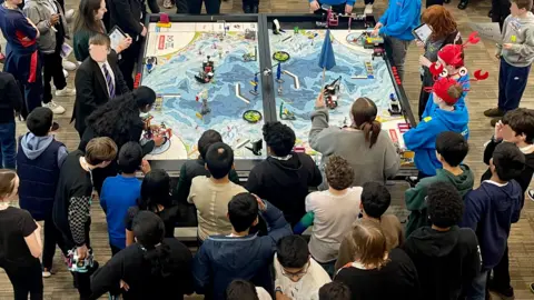 A crowd of approximately 40 children gather around a large table covered by a map. Lego models are sat on the map. A child in the left corner can be seen adjusting one of the models.