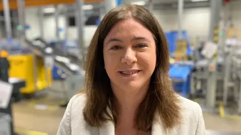 Sam Read/BBC Alex Maher with long brown hair smiling at the camera. She is standing in a factory with shelving and equipment behind her.