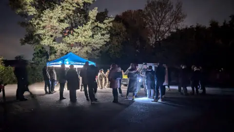 People gathered in a car park at night with members of the military near a blue gazebo with a light. Trees can be seen in the background.