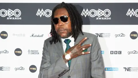 Getty Images Vybz Kartel at the Mobos in Newcastle. He wears a grey suit with a white shirt and green tie, as well as dark sunglasses and a watch. He holds his right arm across his chest, his hand making an 'E' gesture. 