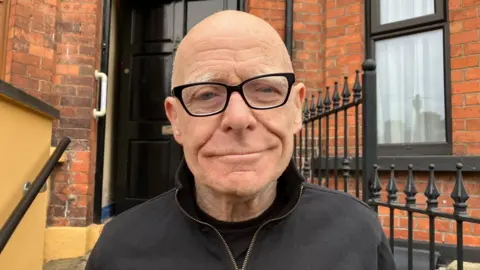 Eamonn McCann stands on steps with railings and the front door a house behind him. He is wearing a black zip up top and black rimmed glasses