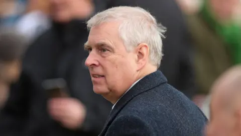 Reuters Prince Andrew stands outside wearing a coat, in this photo from the side with the background blurred. He has white hair and a neutral expression. 