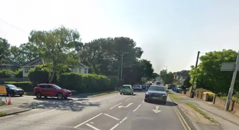 Herbert Avenue, a single carriageway main road with houses on both sides, has a turning lane into Manor Road, seen on the left with cars waiting at a give-way line.