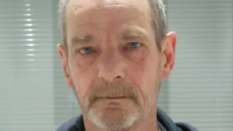 A mugshot of a man with a grey goatee, grey hair and short beard. He has blue eyes and looks into the lens with a neutral expression.