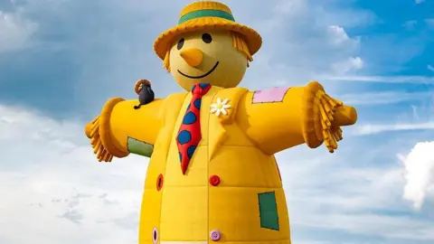 Claire Hartley A large yellow hot-air balloon in the shape of a scarecrow, wearing a red and blue tie and a hat, floating in the air.