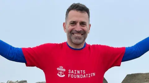 Franny Benali smiling with his arms out as he looks at the camera wearing a Saints foundation t-shirt.