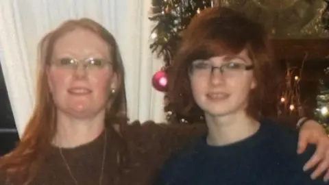 Woman on the left is wearing glasses and has long ginger hair. She is also wearing a brown jumper with a long gold necklace around her neck. She has her arm around a younger woman who is wearing glasses and short ginger hair in a bob and is wearing a blue jumper.