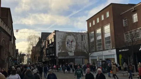 Exeter High Street with a mural showing a woman's face , shops and the street busy with shoppers 