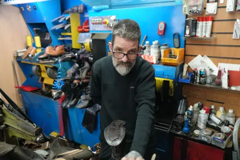 BBC news A man looks at the camera. He is wearing dark clothes and glasses. He is surrounded by shoes and equipment to fix them. 