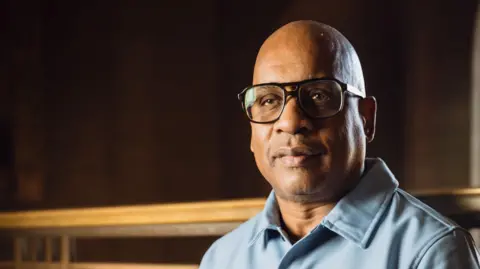 Glenn Ligon wearing large framed dark glasses and a light blue shirt, staring at the camera.