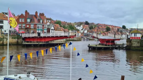 Whitby swing bridge opening with part of Whitby behind it and some blue and yellow bunting.