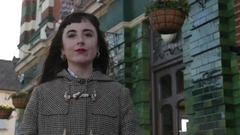 Hayley standing outside the Painters Arms in High Town. She has long straight brown/reddish hair with a short fringe, wears a nose ring and has bright red lipstick on.