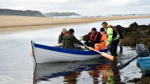 Three people sit in a white and blue rowing boat and are helped out into an inlet by three others. Waves crash on a beach behind them.