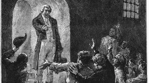 Getty Images Black and white drawing of a man in light-coloured clothes  and a black jacket. He is standing in front of a group of prisoners, some of whom have their hands raised. He is pointing in their direction while holding a top hat with the other hand.